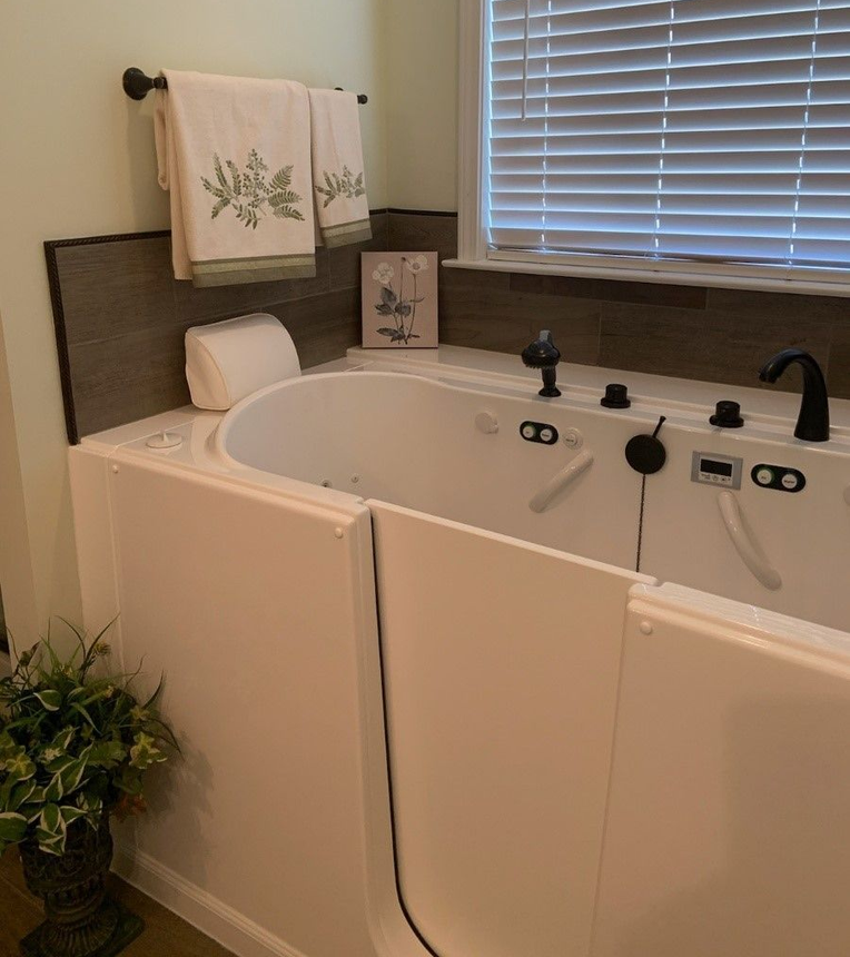 White walk-in bathtub with a side door, controls, and faucet, installed in a bathroom with towels and a window.