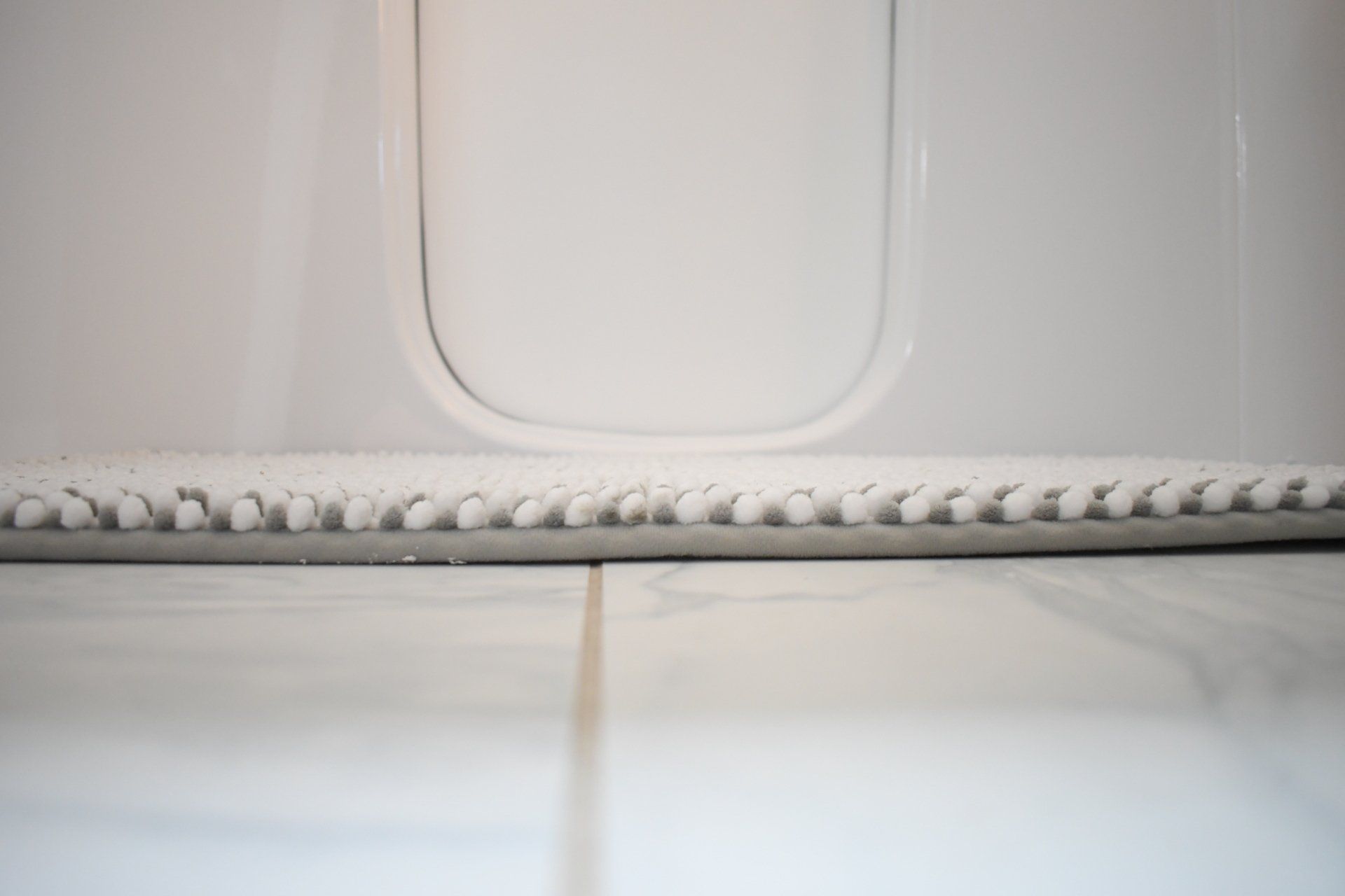 A low-angle view of a gray-backed, white bath mat resting on a white tiled floor against a bathtub.