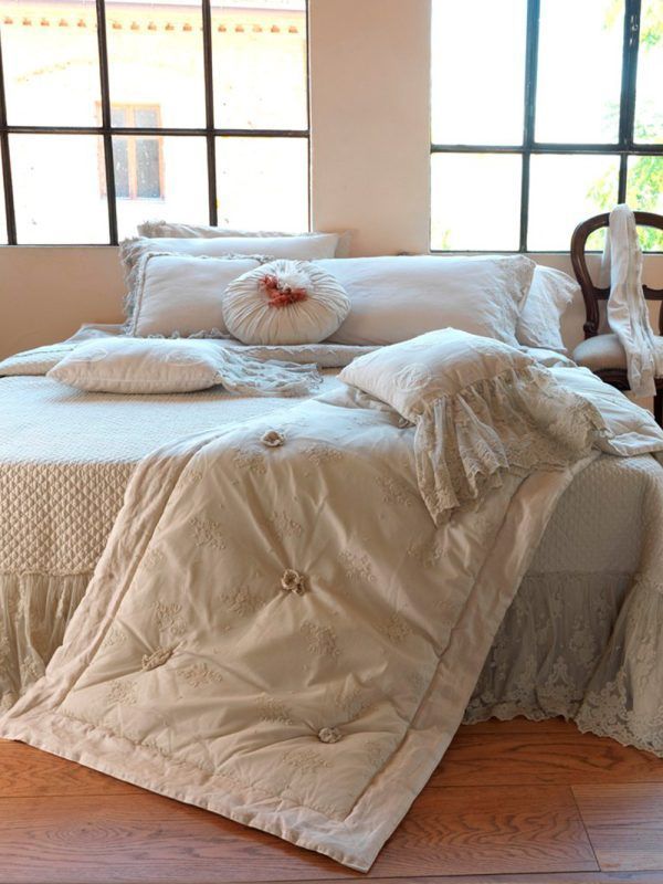 Letto con biancheria da letto di colore chiaro, tra cui una trapunta e cuscini arricciati, vicino a una finestra.