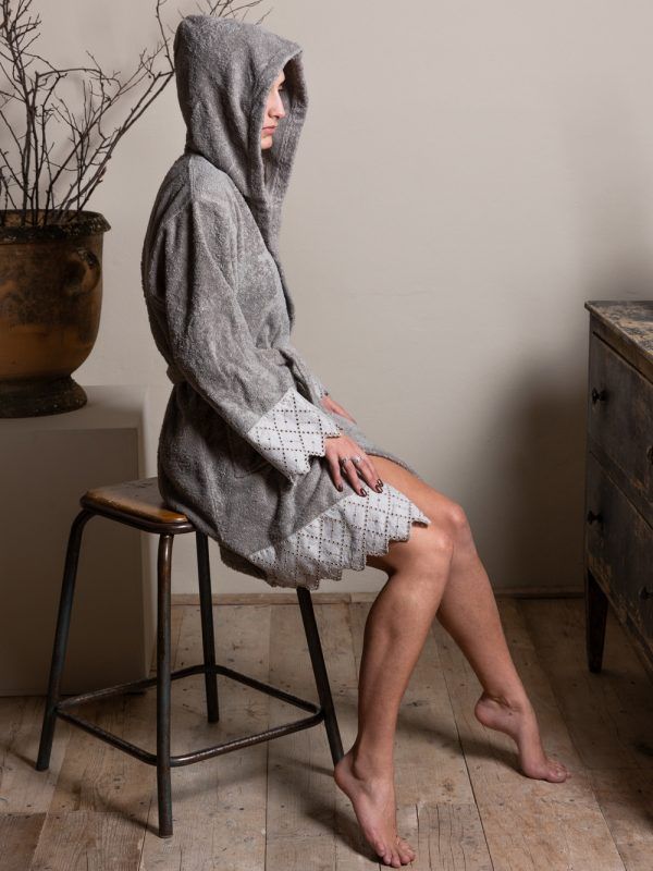 Donna in tunica grigia con cappuccio, seduta su uno sgabello, a piedi nudi. Maniche e orlo decorati in pizzo.
