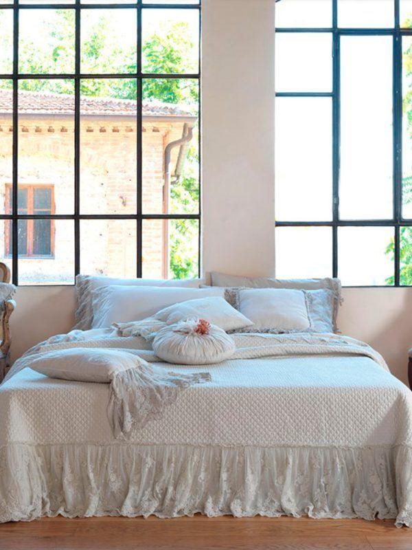Letto con lenzuola bianche, bordi in pizzo e cuscini decorativi di fronte a grandi finestre.