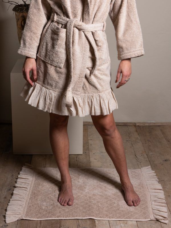 Donna che indossa una vestaglia beige con orlo arricciato, in piedi su un tappetino da bagno coordinato in una stanza.