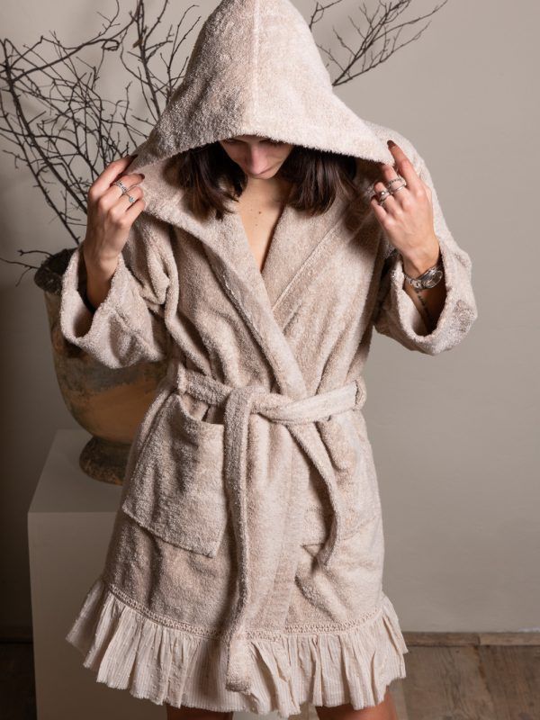 Donna che indossa un accappatoio beige con cappuccio e cintura, in piedi vicino a un vaso con rami.