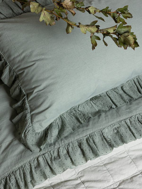 Cuscino verde salvia con bordo arricciato su un letto, decorato con un ramo frondoso.