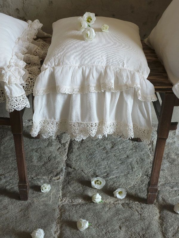 Panca bianca con gonna in tessuto arricciato e cuscini, decorata con fiori bianchi su un pavimento in pietra.