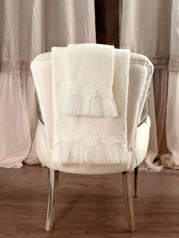 Asciugamani color crema con bordi in pizzo sono drappeggiati sullo schienale di una sedia bianca decorata, davanti a tende arricciate.