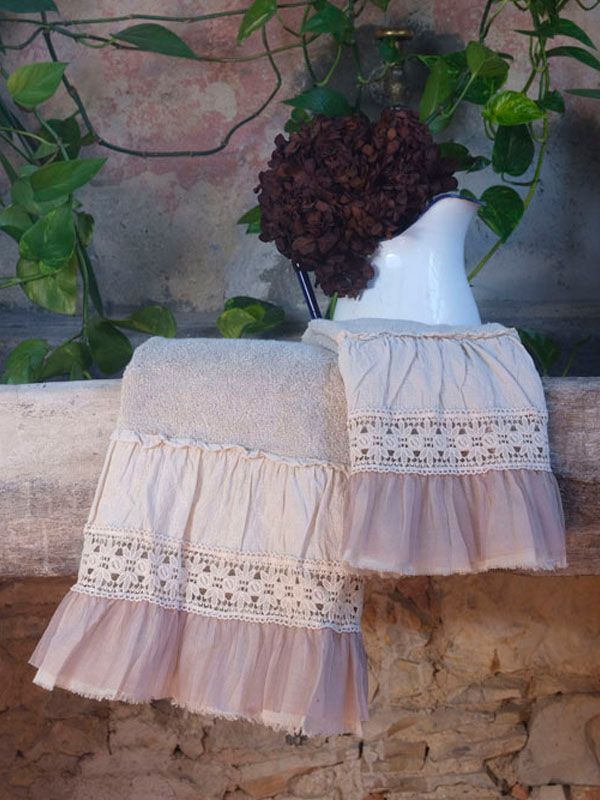 Due asciugamani, uno color crema e uno rosa, con bordi in pizzo, sono drappeggiati su una rustica mensola in pietra. Una brocca bianca contiene fiori secchi.