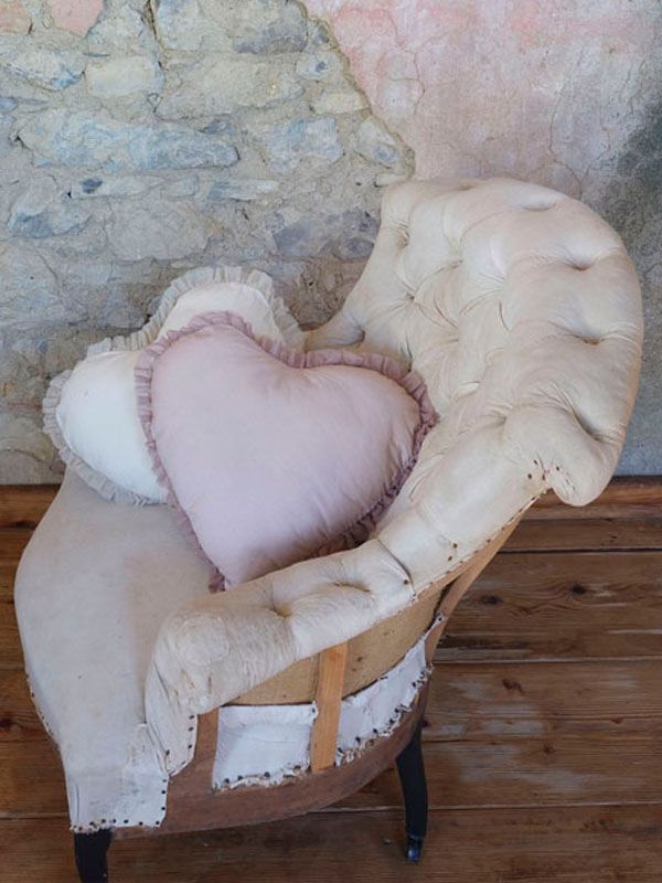 Una poltrona color bianco sporco e consumata, con schienale capitonné e due cuscini a forma di cuore, contro una parete strutturata.