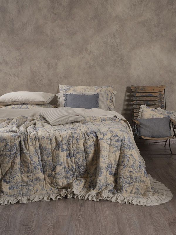 Letto con trapunta floreale blu e cuscini coordinati, sedia in legno, contro una parete grigia testurizzata.