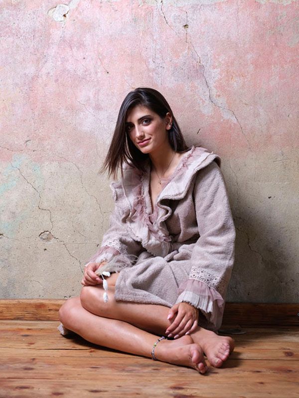 Una donna con una tunica beige è seduta a gambe incrociate su un pavimento di legno, contro una parete ruvida nei toni del rosa.