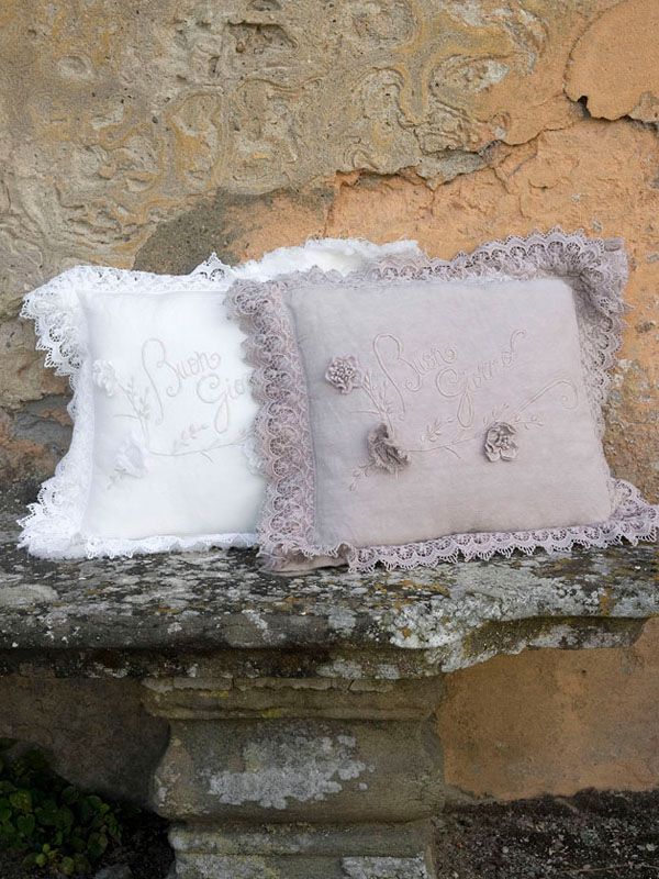 Due cuscini decorativi quadrati con bordi in pizzo, bianchi e viola pallido, su una mensola in pietra.