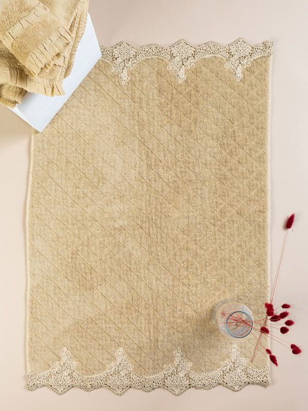 Runner da tavolo rettangolare beige con bordi in pizzo, adagiato su una superficie rosa chiaro, con asciugamani piegati e un vaso con fiori rossi.