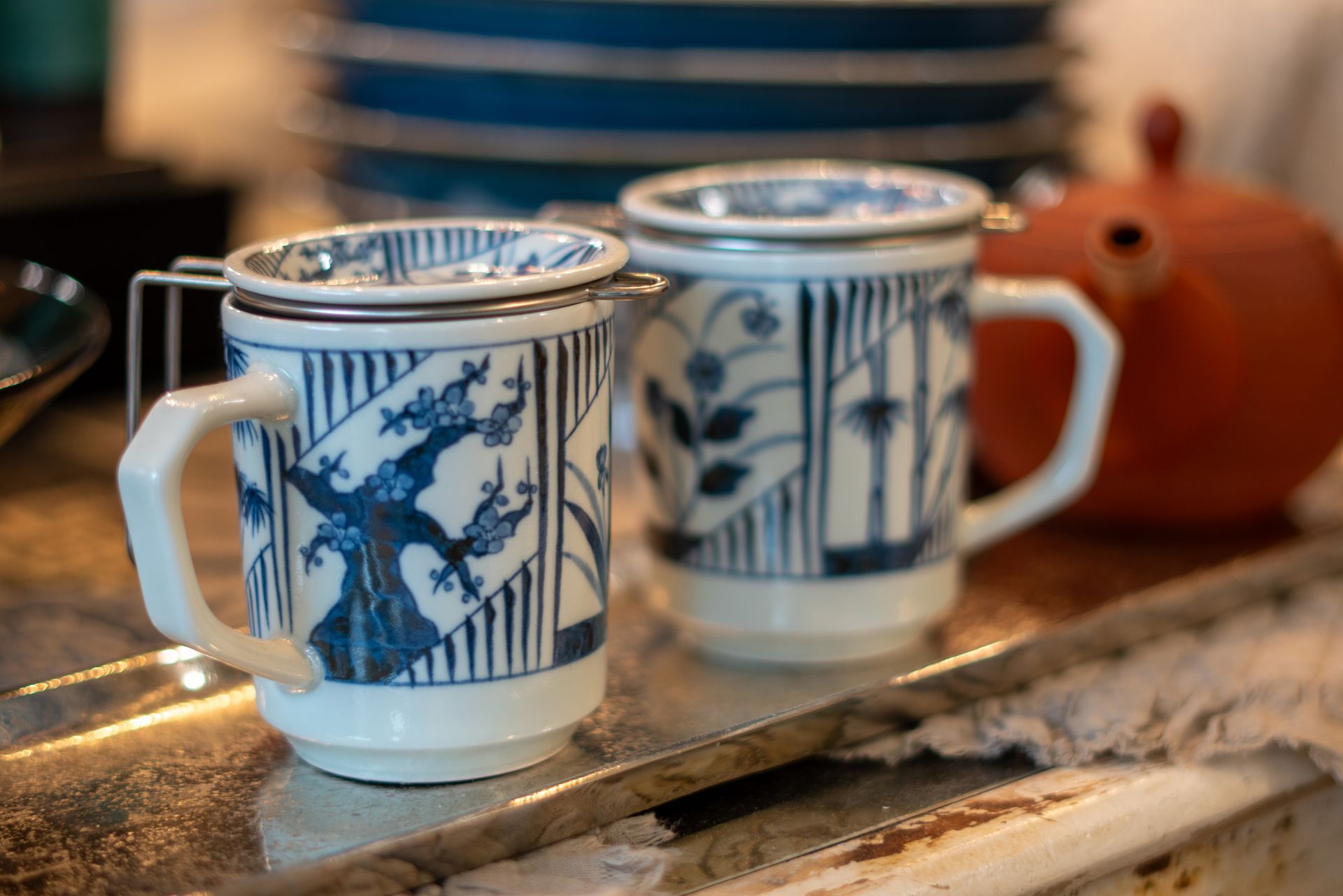 Due tazze da tè con decori blu e bianchi, dotate di colini, appoggiate su una superficie riflettente, con piatti impilati e una teiera sullo sfondo.