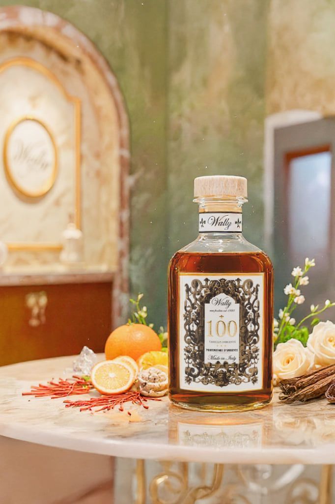 Una bottiglia di liquido ambrato con etichetta è appoggiata su un tavolo di marmo con frutta e fiori, su uno sfondo decorato.