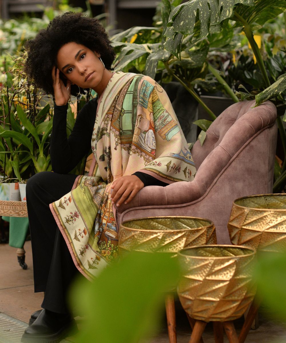Donna vestita di nero con una sciarpa fantasia, seduta su una sedia rosa tra le piante, con vasi dorati in primo piano.
