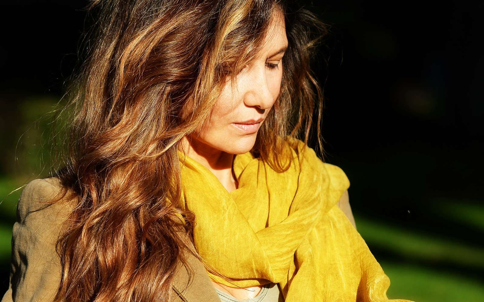 Donna con lunghi capelli castani, che indossa una sciarpa dorata e una giacca marrone, guarda verso il basso, all'aperto.