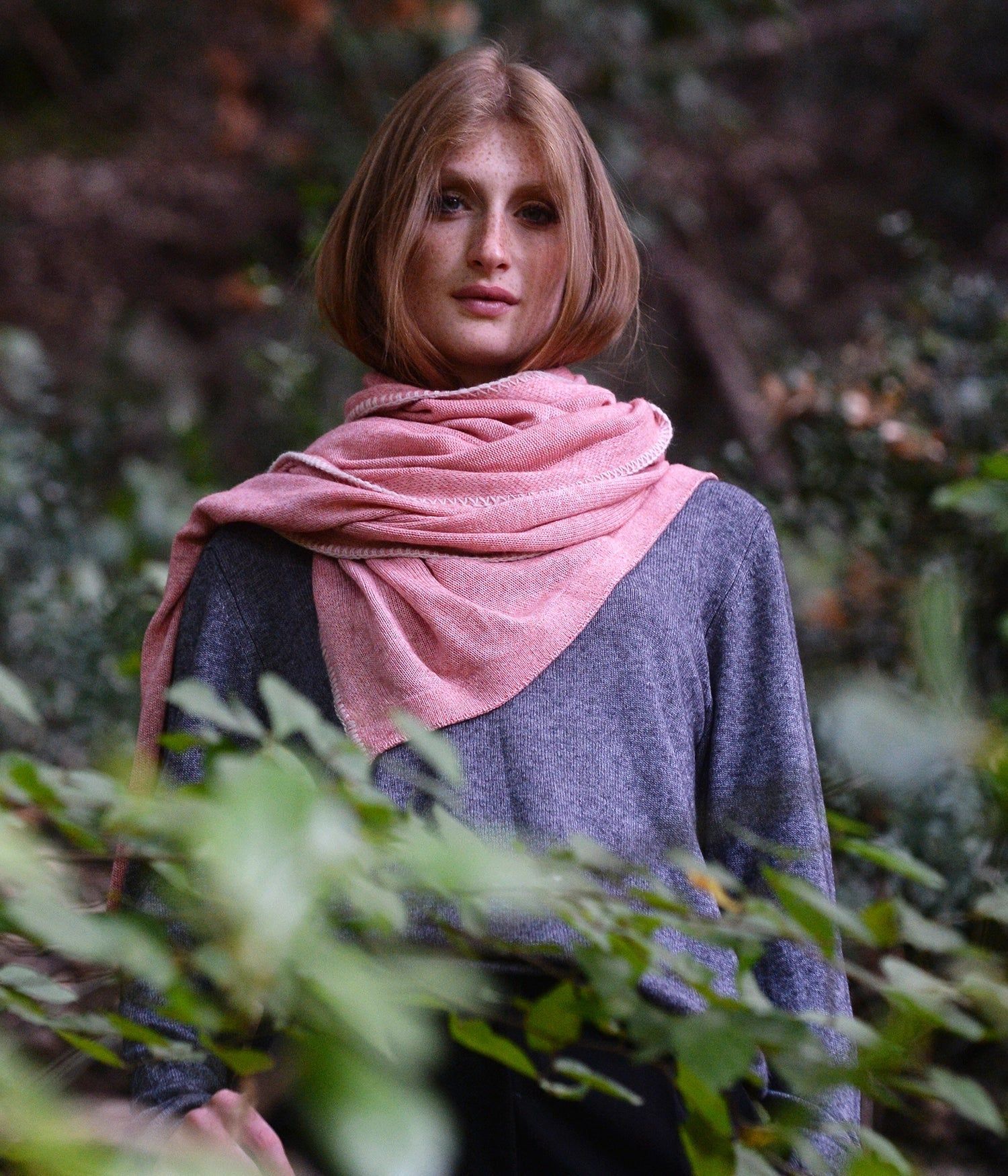 Donna con maglione grigio e sciarpa rosa in un ambiente boscoso.