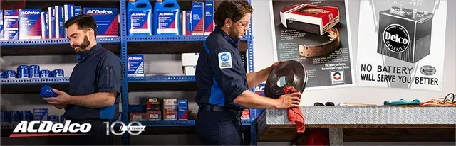 Two mechanics in a shop, one examining a part, the other holding a helmet, with product advertisement posters visible.