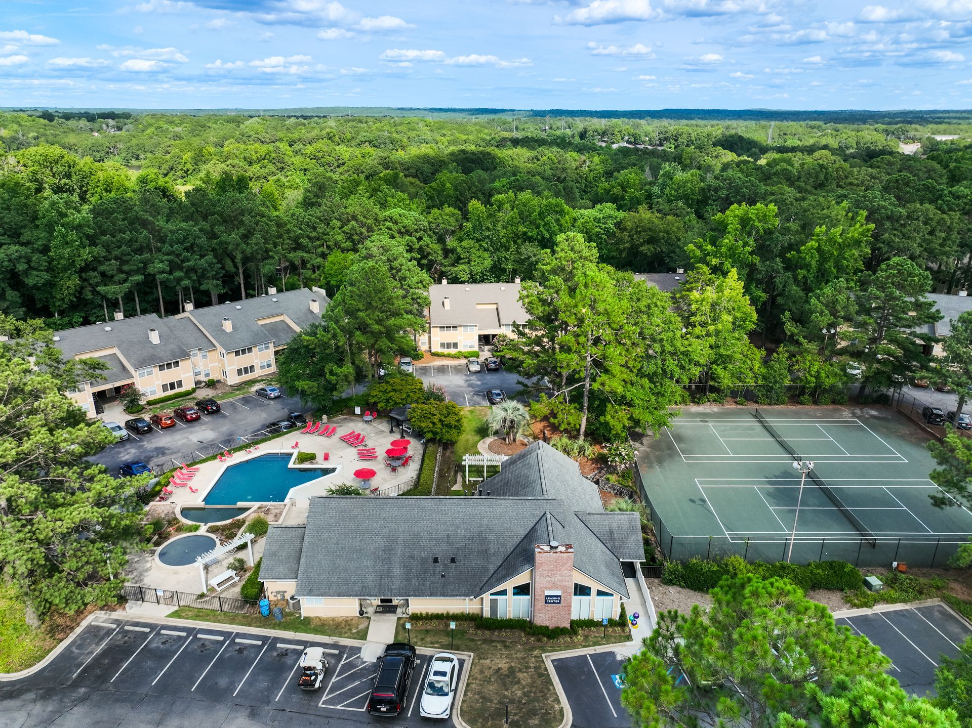Aerial view of a residential complex with a pool, tennis courts, and buildings surrounded by trees.
