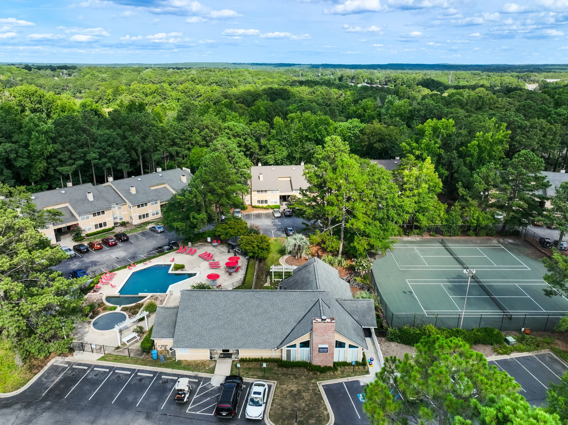 Aerial view of a residential complex with a pool, tennis courts, and buildings surrounded by trees.