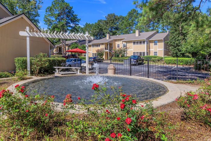 Fountain in a landscaped area with red roses, a bench, and apartment buildings in the background.