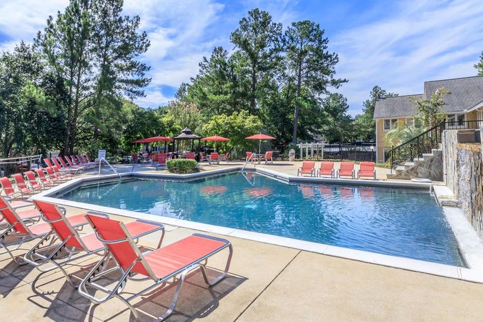 Pool area with red lounge chairs, clear blue water, and trees under a bright sky.