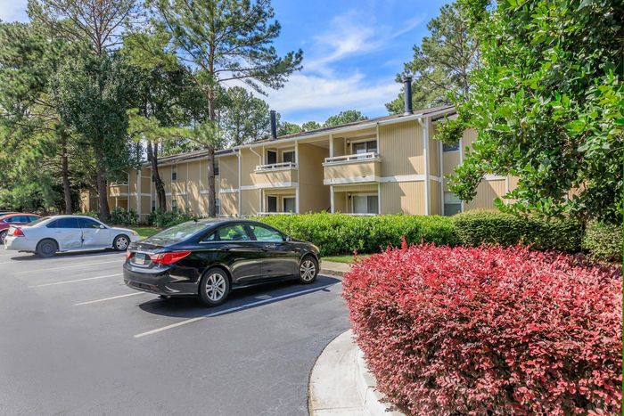 Apartment building with tan siding, balconies, and parked cars in a parking lot with a red bush in front.