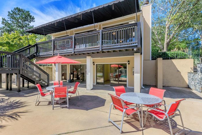 Two-story building with patio, red tables/chairs. Stairs lead to upper deck. Blue sky, trees in background.