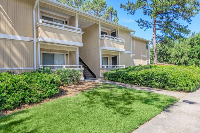 Apartment building with beige siding, balconies, and lush green landscaping under a blue sky.