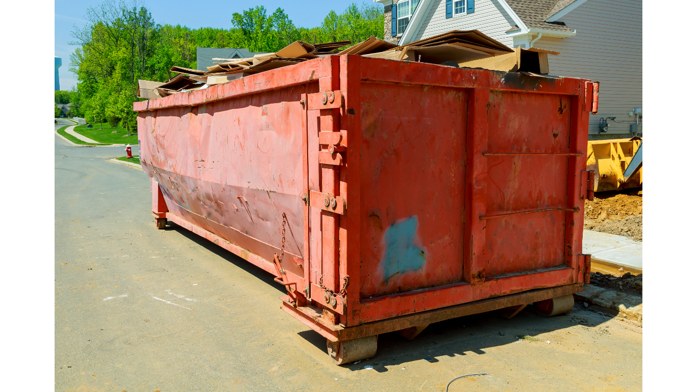 Red construction dumpster on a paved street, filled with debris. A house and trees are in the background.