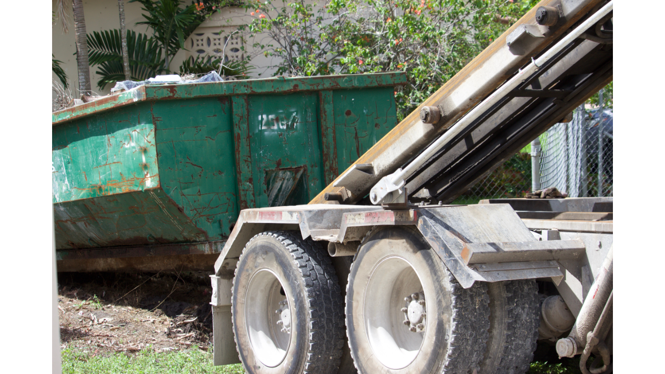A green dumpster being lifted by a truck with large tires. The dumpster is full of trash.