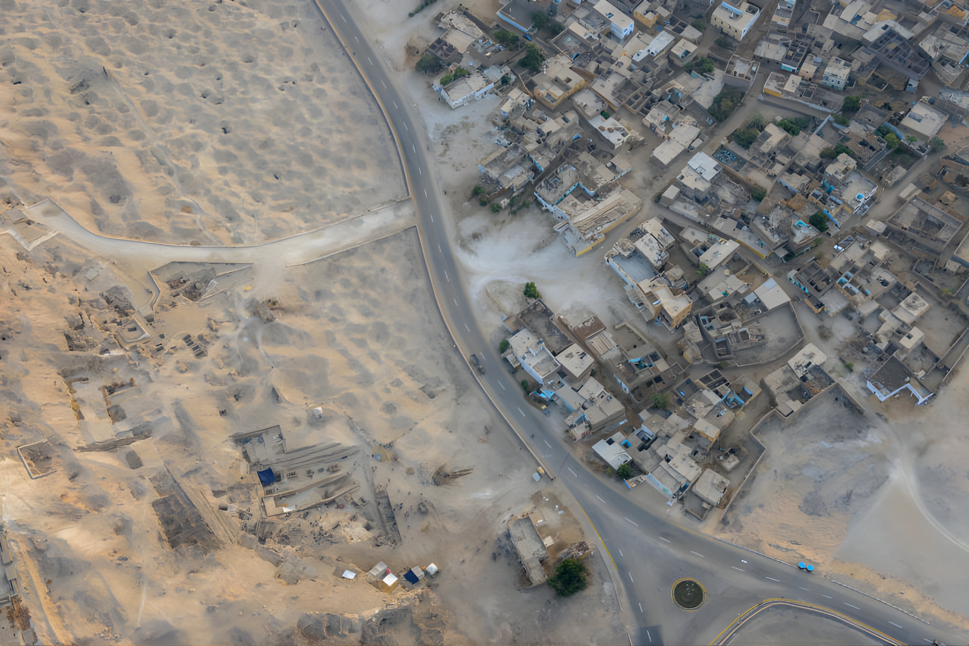 Aerial view of a town next to a desert. Road curves through the scene, buildings densely packed.