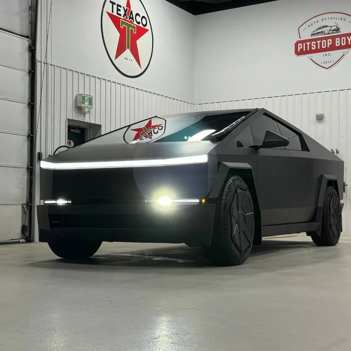 Matte black Tesla Cybertruck in a garage with headlights on, viewed from the front-left.