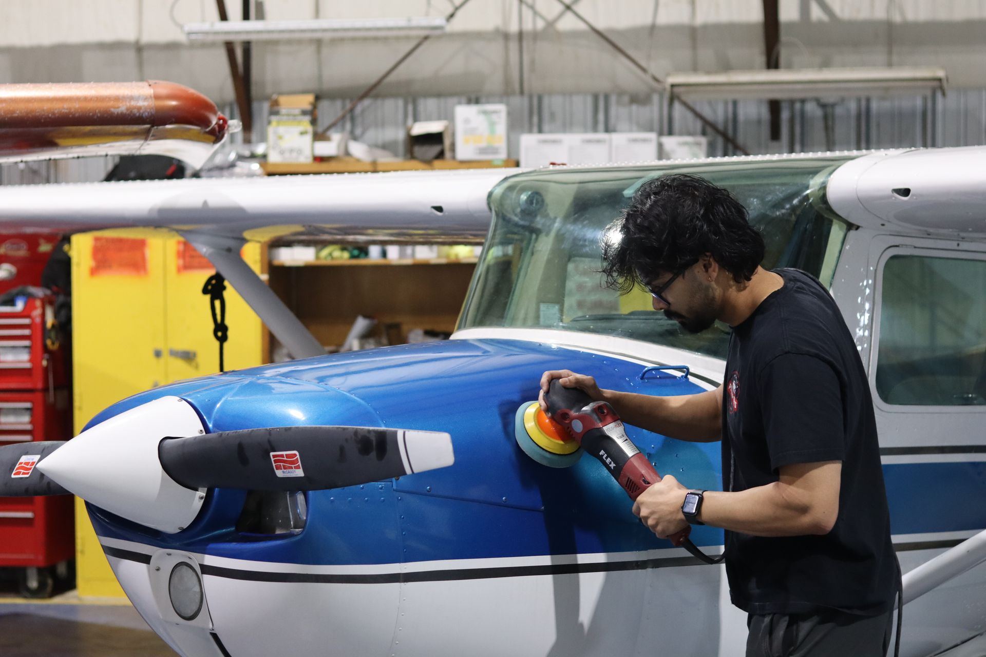 Small blue-and-white propeller airplane parked in a hangar near an orange equipment cabinet.