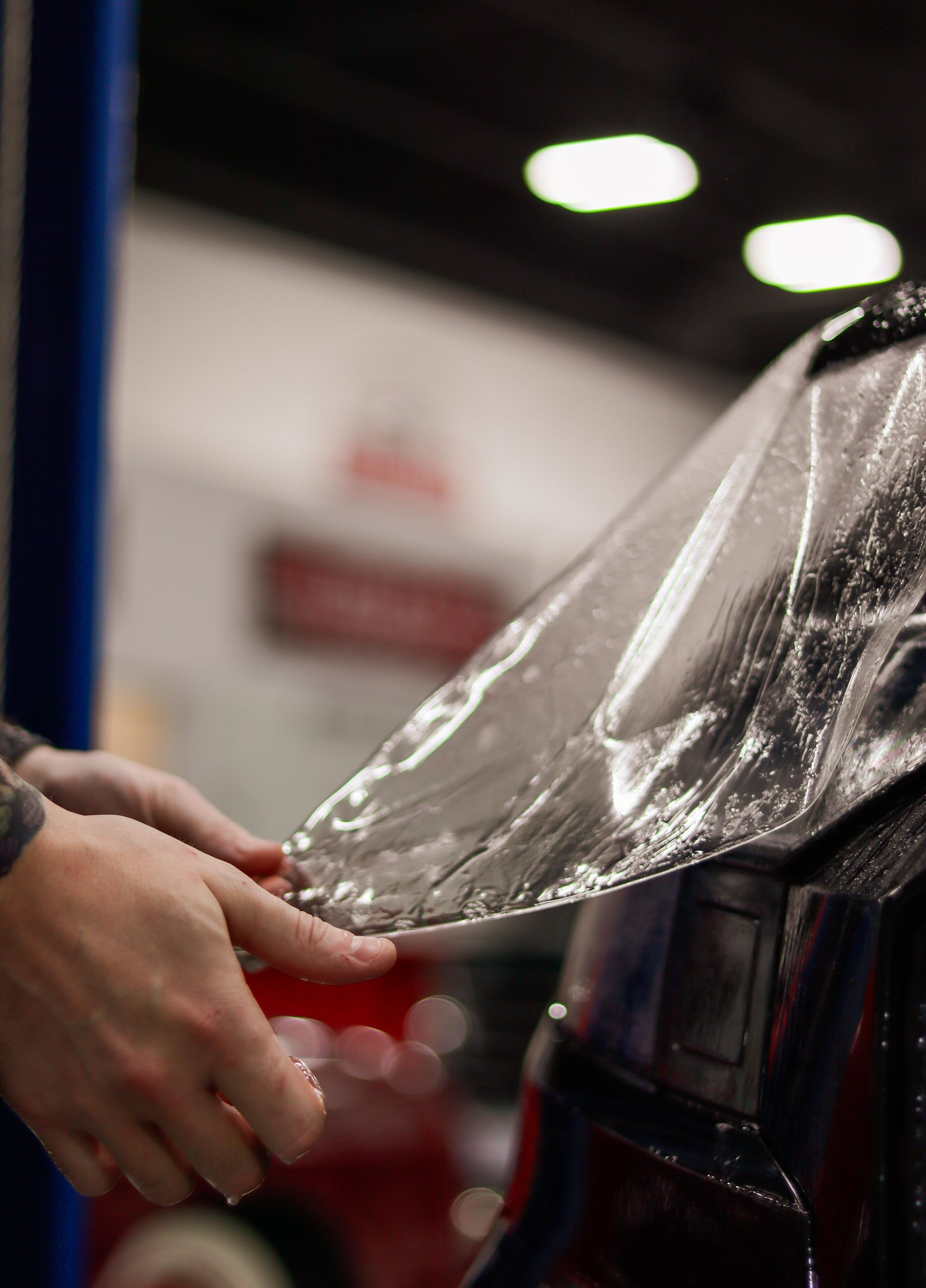 Hands peeling clear protective film from a dark car surface at an indoor auto show.