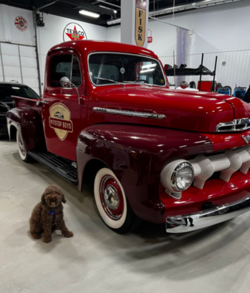 Red vintage pickup truck in a garage, with a small brown dog sitting beside it.