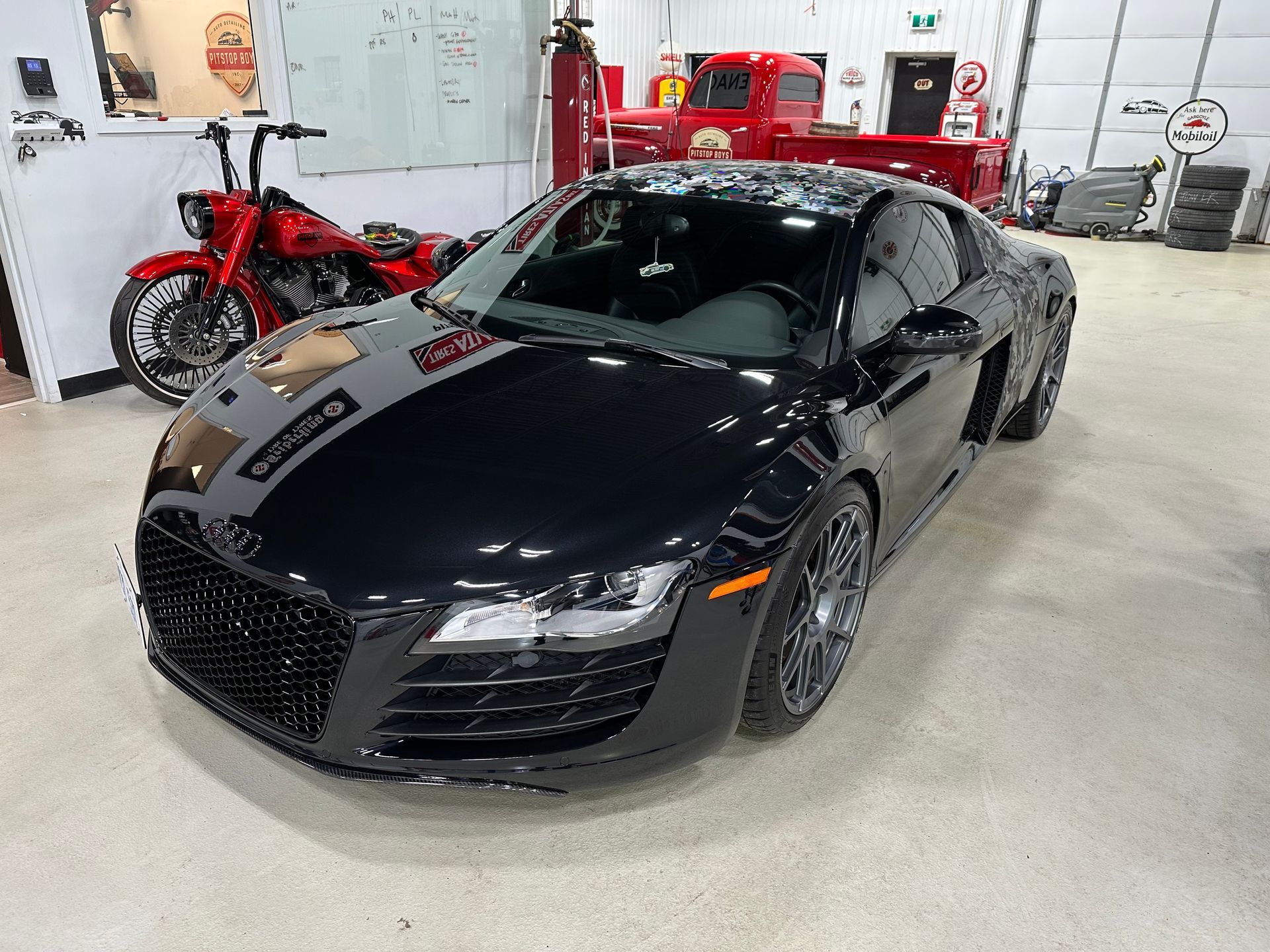 Black Audi R8 in a garage showroom with red motorcycles and service equipment in the background