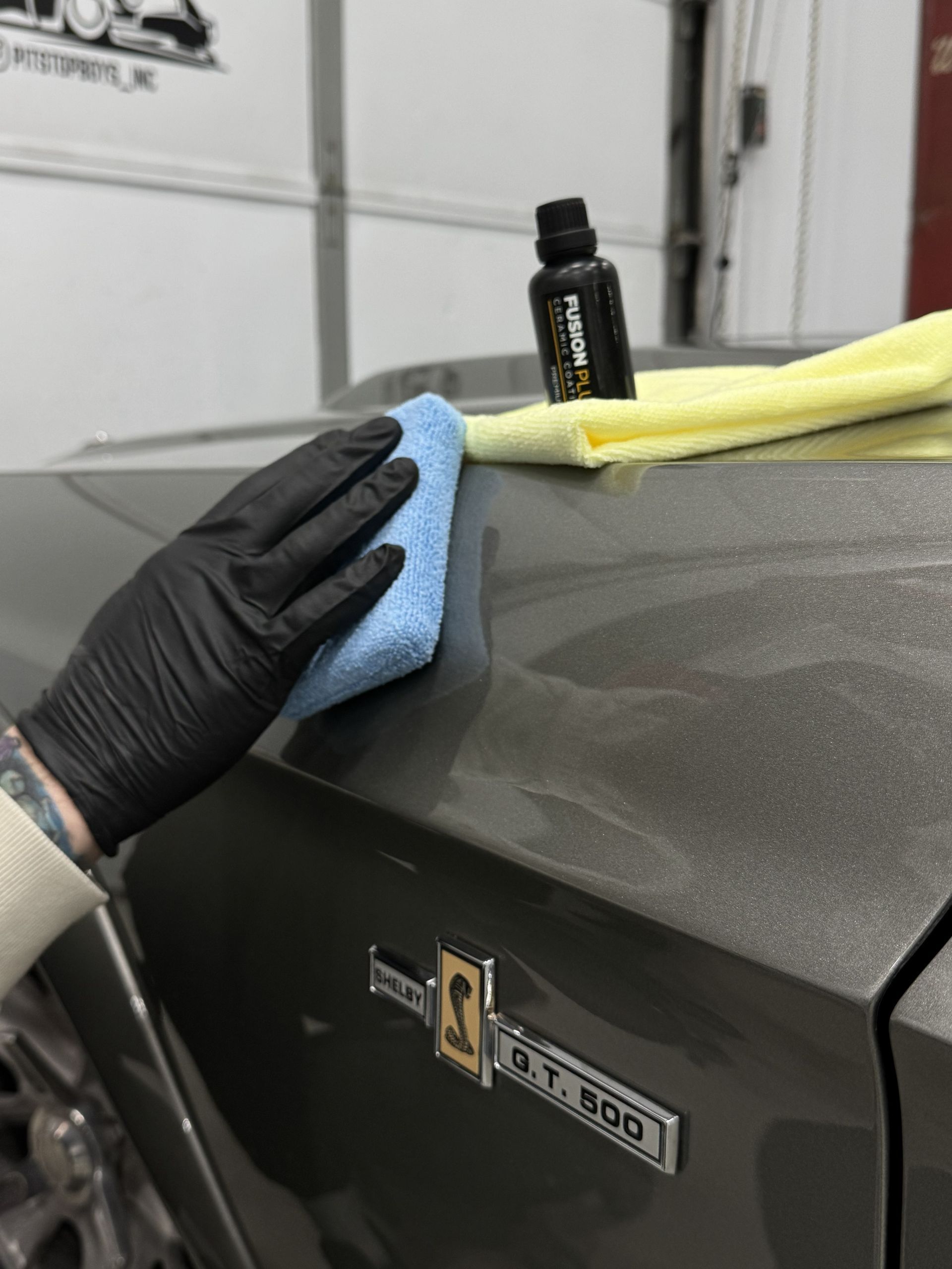 Black-gloved hand wiping a dark car hood with a blue cloth in a garage.