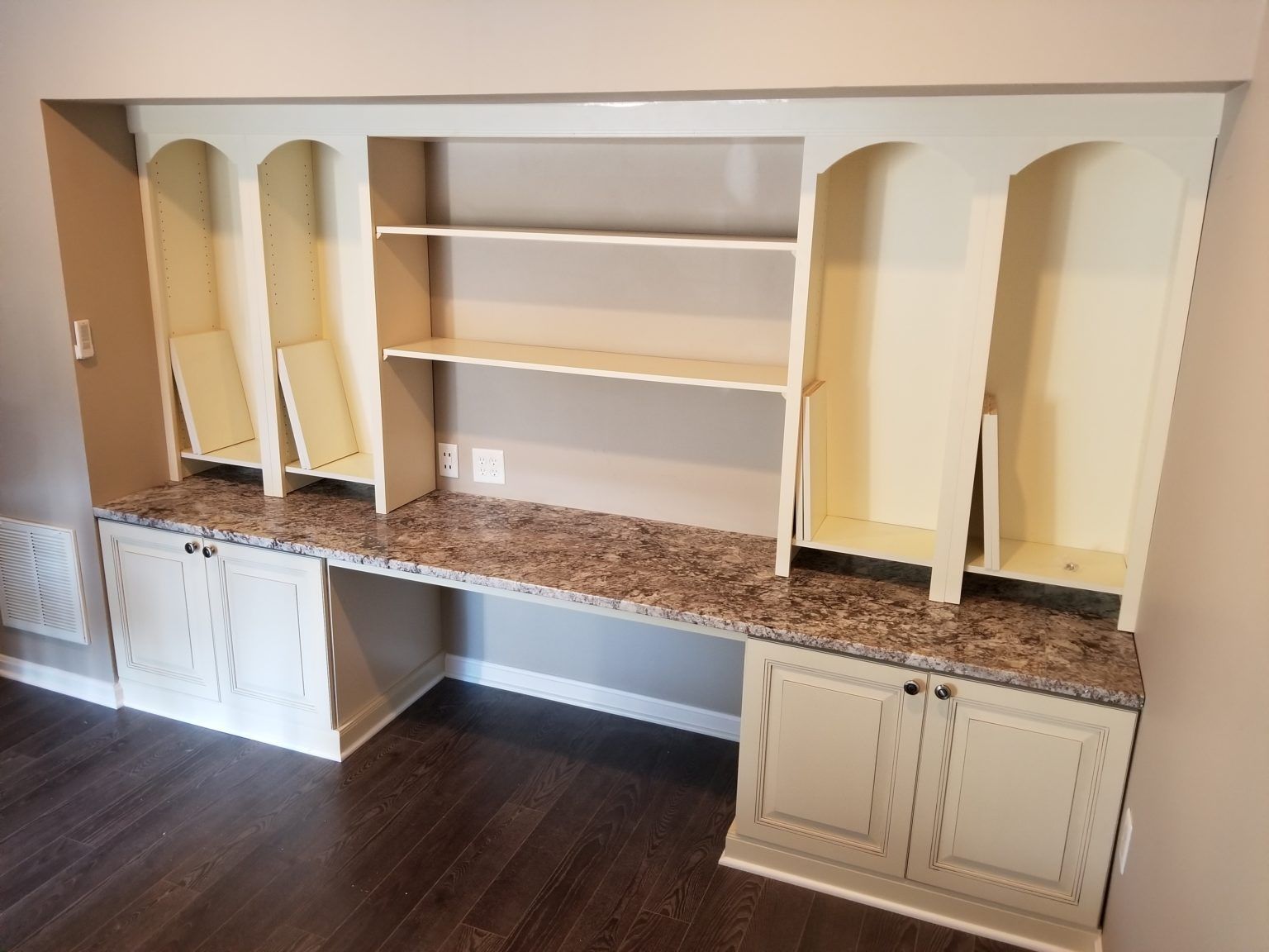 Built-in beige desk with upper shelves and cabinets, against a light brown wall.