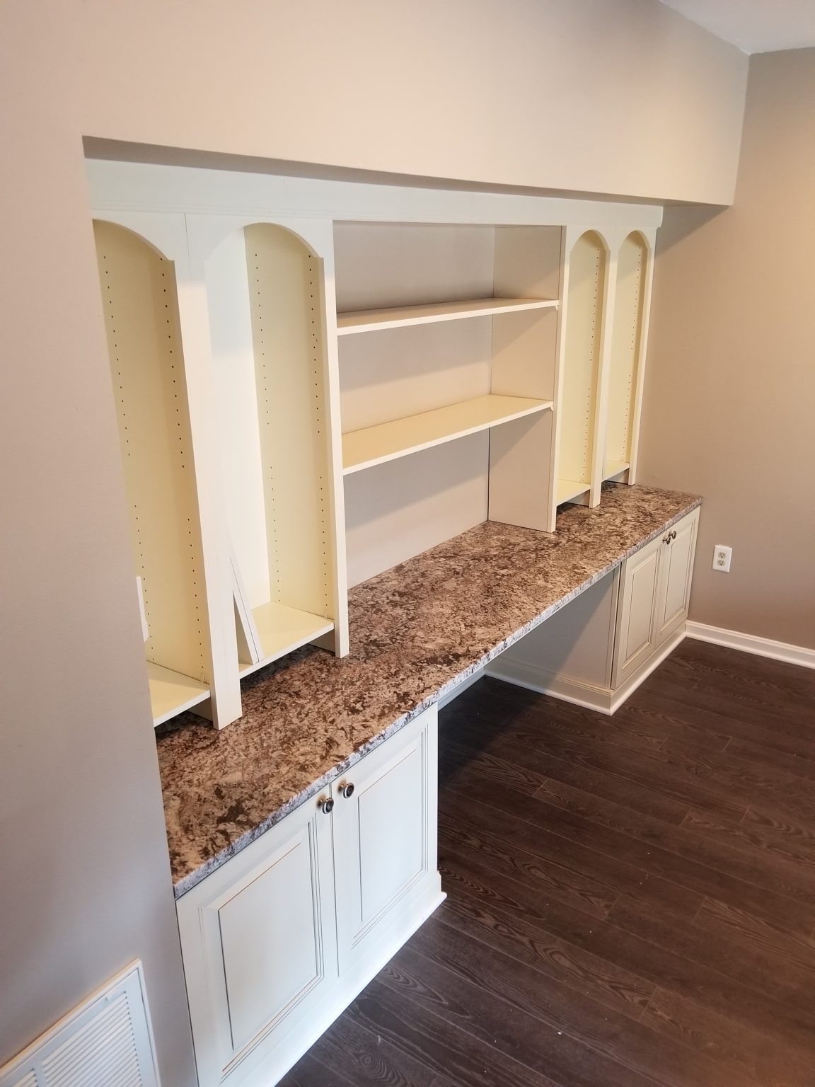 Built-in white desk with shelves and granite-like countertop in a beige-walled room with dark wood flooring.