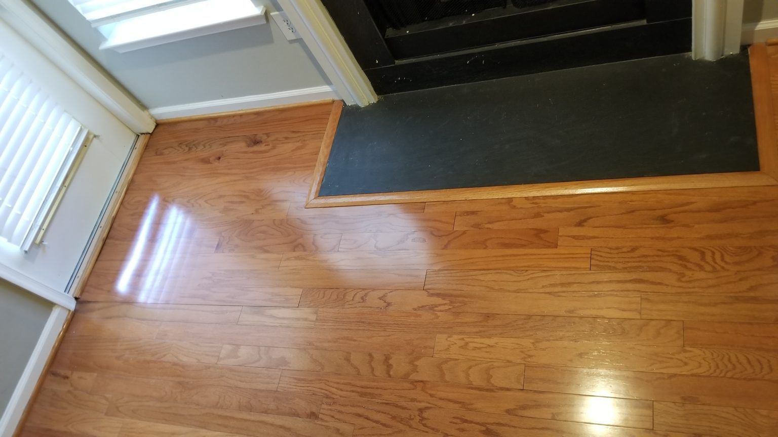 Hardwood floor, doorway, and black doormat. Sunlight reflecting on the wood.