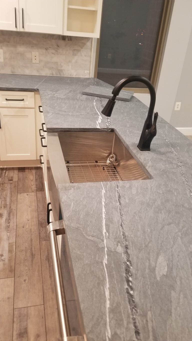 Kitchen countertop with sink and faucet, gray stone with white grout, dark cabinet handles.
