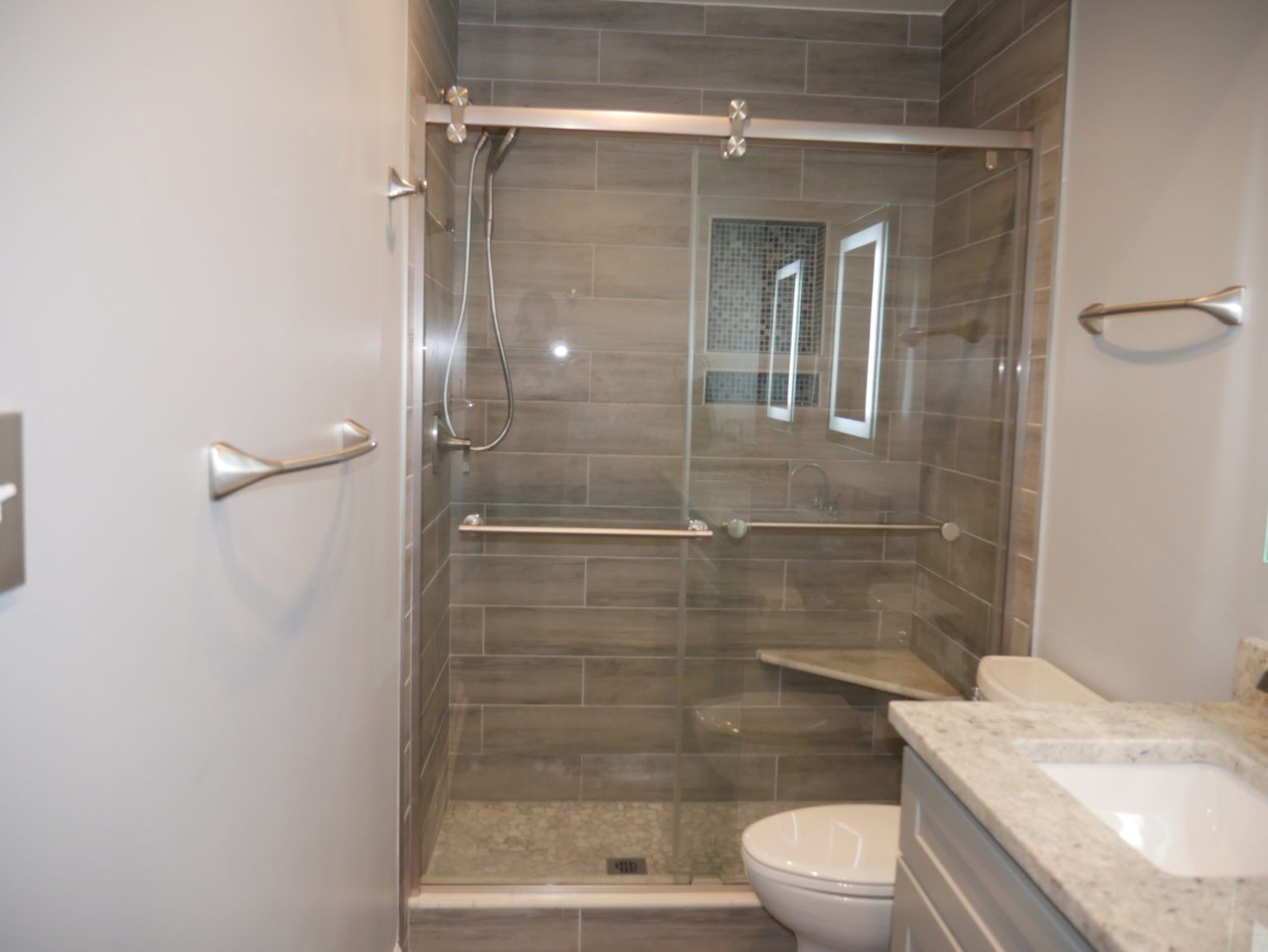Modern bathroom with gray tiled shower and glass door. Toilet and vanity visible.