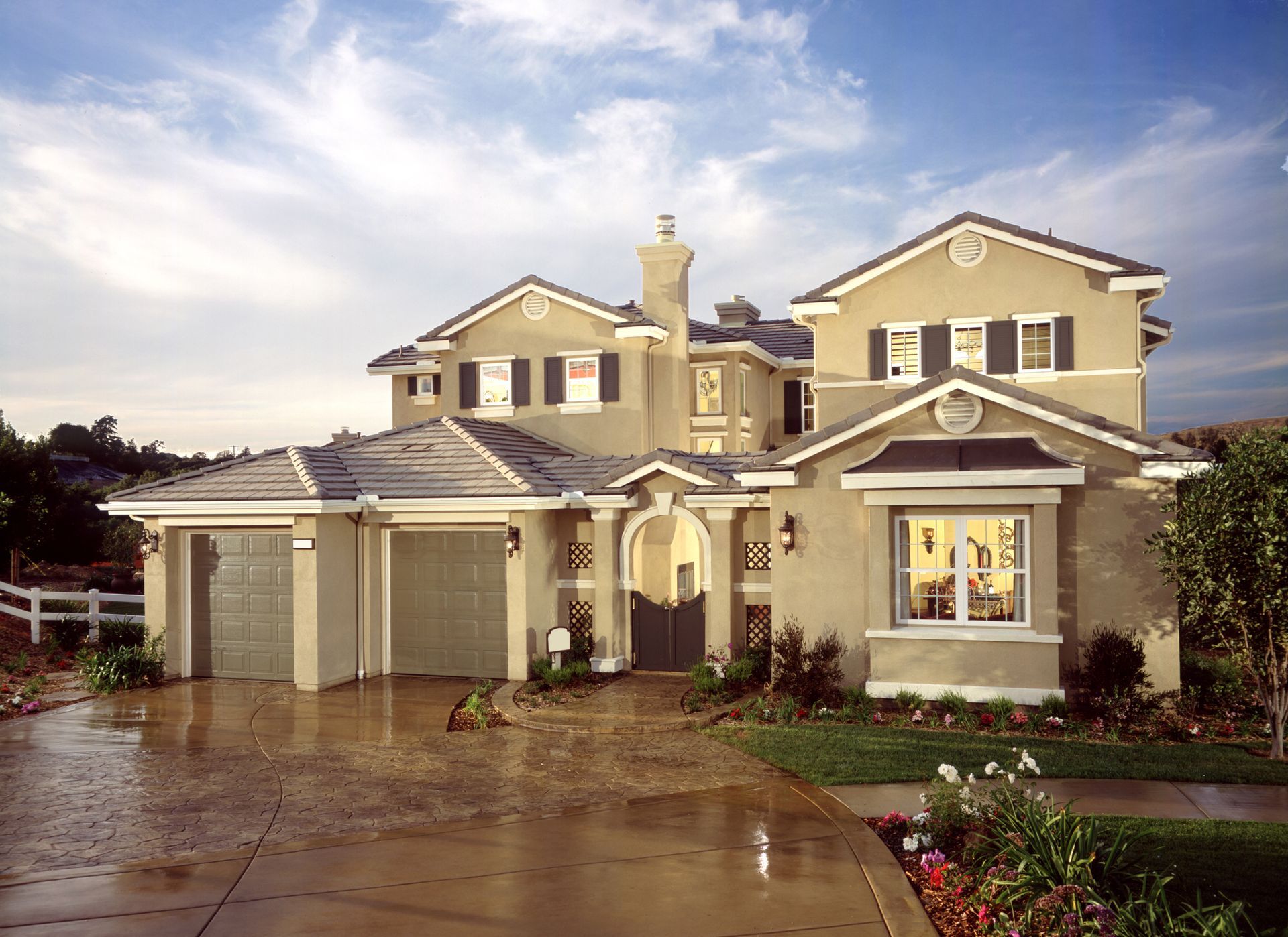 Two-story beige house with a three-car garage, driveway, and landscaping under a cloudy sky.
