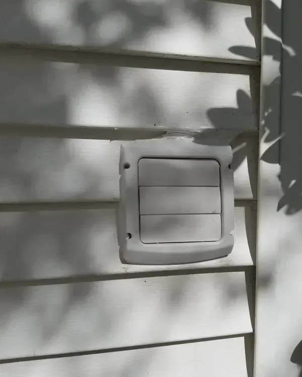 White dryer vent on white siding, with tree shadows.