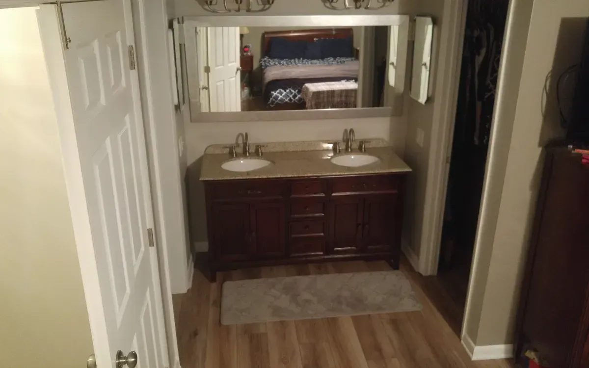 Bathroom with double vanity, mirror, and a glimpse into a bedroom. The vanity is brown, and the floor is wood.