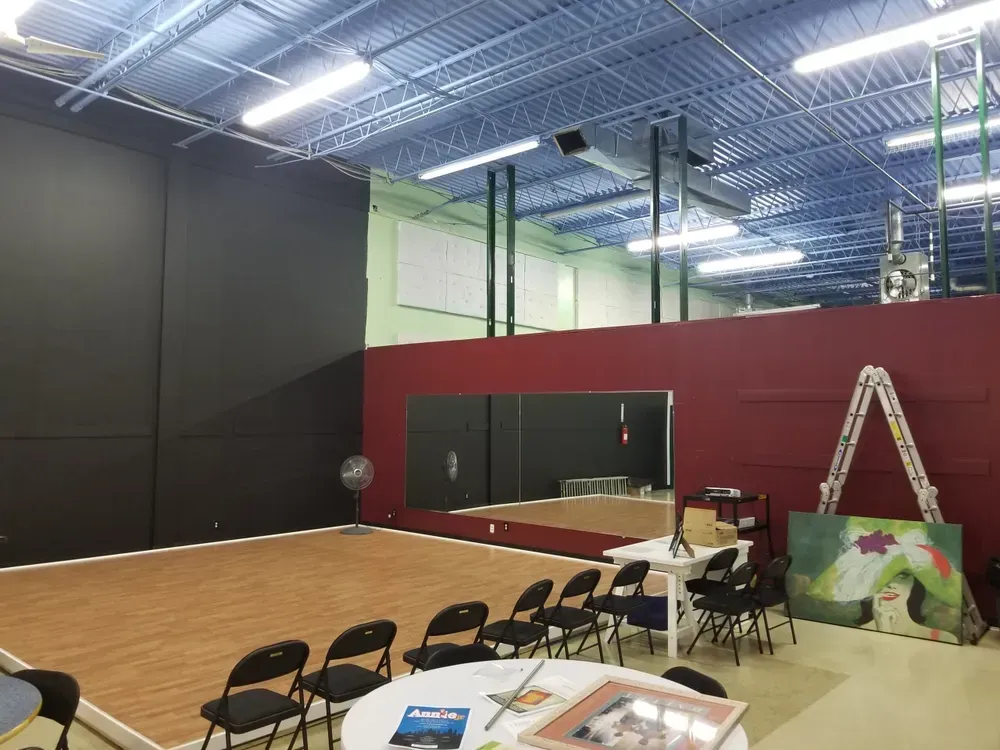 Dance studio with wood floor, mirrors, black wall, chairs, and artwork. Blue ceiling.