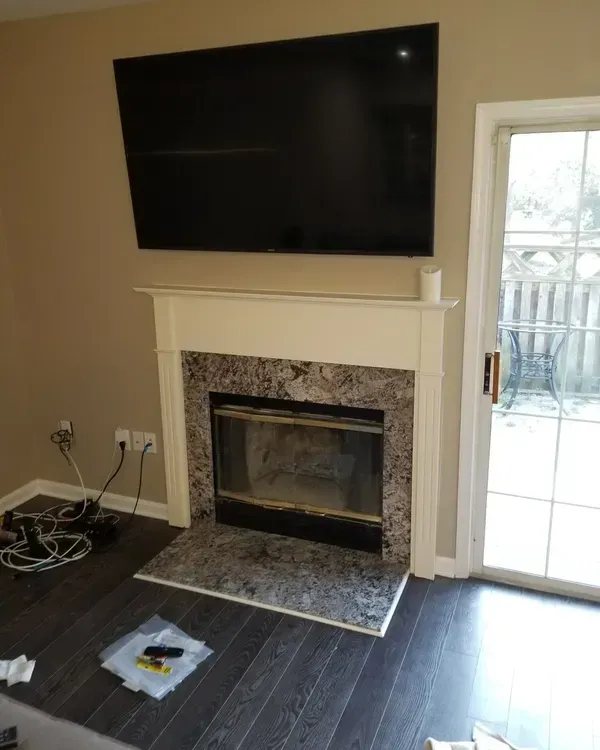 Black TV above a fireplace with a granite face. Dark wood floor, beige walls. Sliding glass door to a patio.