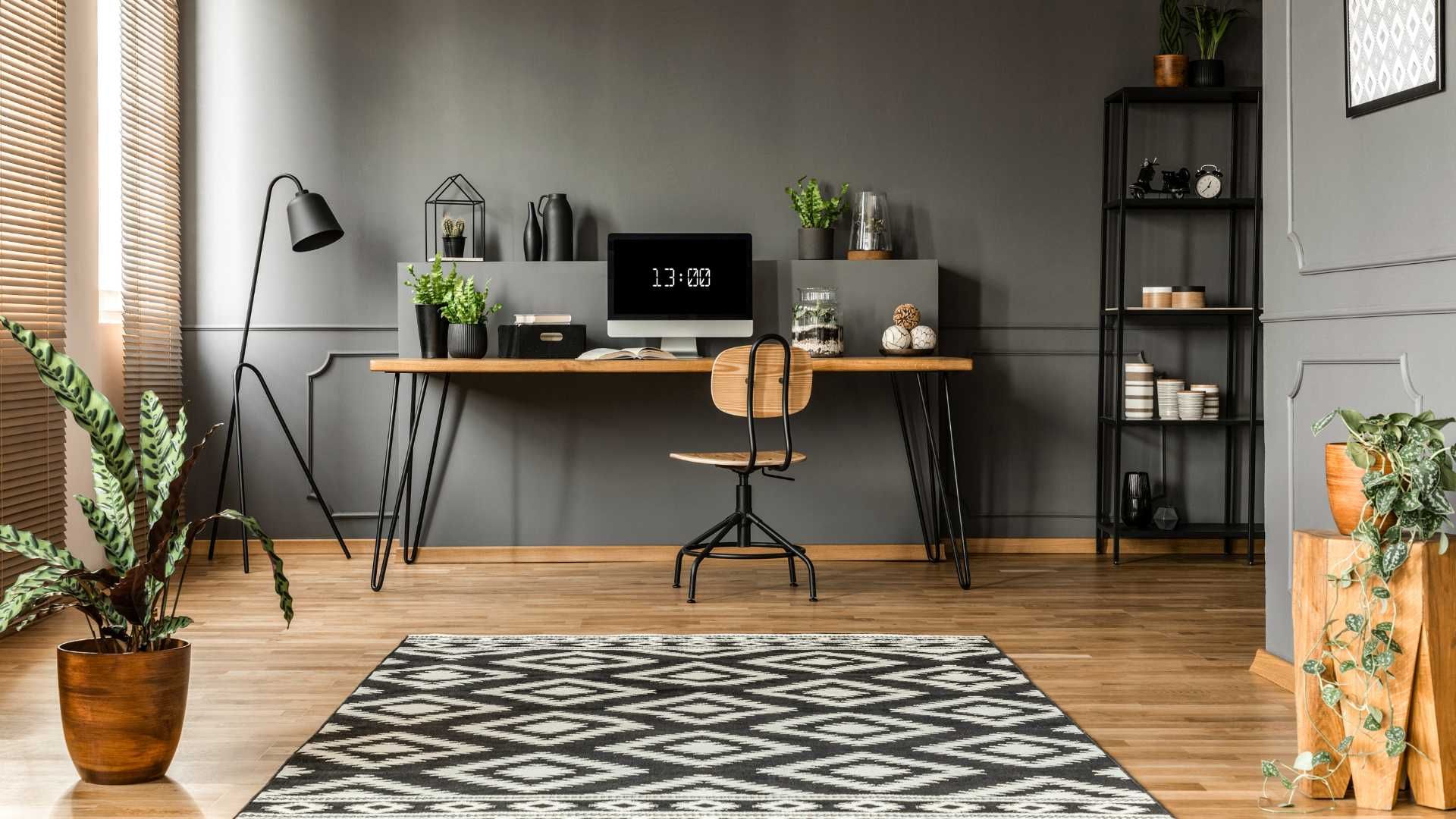 Desk with computer and chair in a gray room. Black patterned rug, plants, and metal shelving visible.