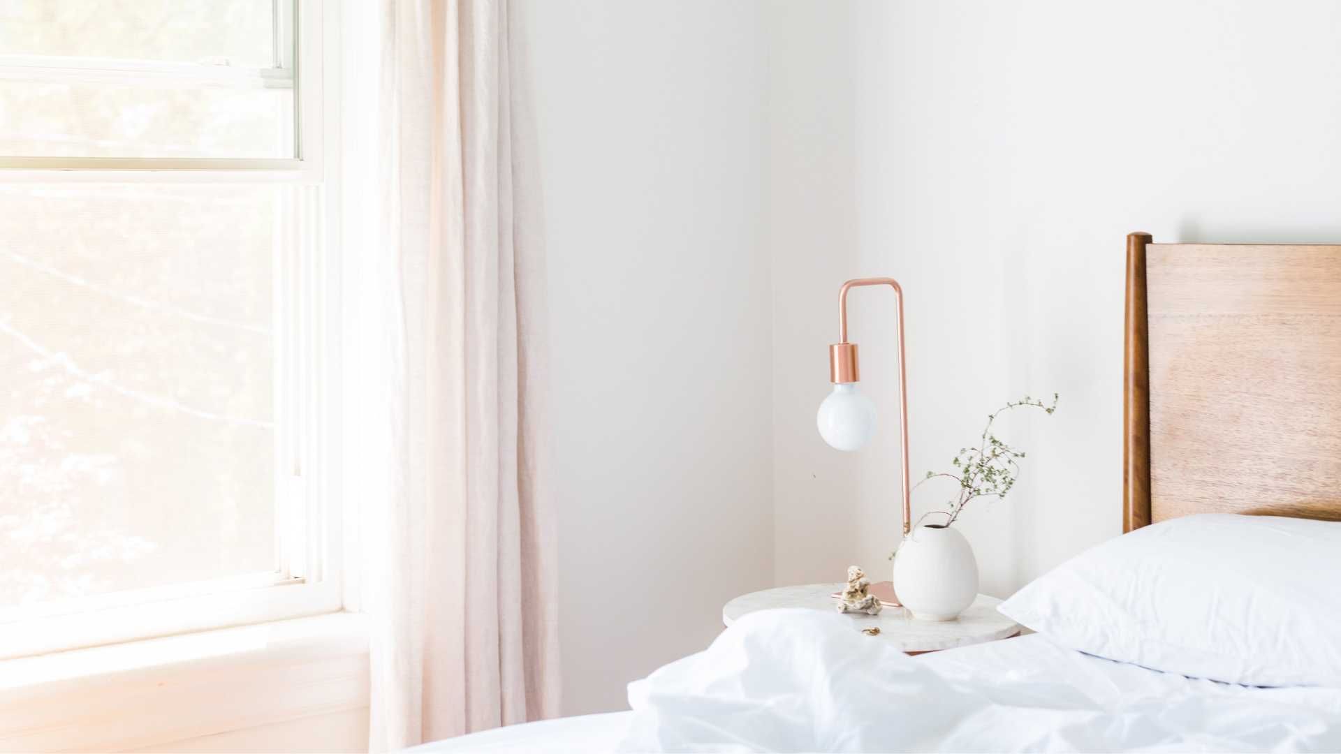 Bedroom with wooden headboard, white bedding, copper lamp, and soft, natural light from a window with curtains.